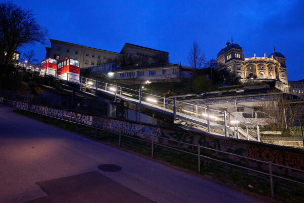 Blick auf die Marzilibahn bei Dämmerung. Von Westen her, 20m von Geleisen entfernt, zeichnet die Beleuchtung entlang der Bahnstrecke eine Linie zur Bergstation, halb verdeckt links oben im Bild. Die rechte obere Hälfte des Bildes zeigt das beleuchtete Bundeshaus unter einem dunkelblauen, bedecken Himmel.