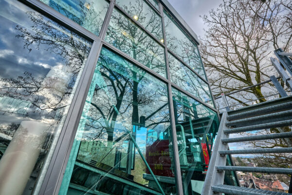 Blick von der Treppe der Marzilibahn Bergstation in Bern. Zwei drittel des Bildes nimmt links die Glasfassade der Bergstation ein. Es spiegelt sich der bewölkte Himmel in den Scheiben. Durch die Scheiben hindurch ist ein roter Wagen der gerade abfahrenden Bahn zu sehen.