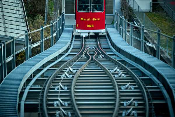 Die strecke der Marzilibahn vom oberen Wagen gesehen. Die Geleise gehen in der Mitte der Strecke symmetrisch auseinander und wieder zusammen. Die Rote Kabine des eben erst losgefahrenen unteren Wagens hebt sich ab vom kühlen, blau-grau gehaltenen Ton des Bildes.